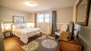 A bedroom with a bed, bedside lamps, a desk with a computer, a chair, a window with curtains, and a motivational quote on the wall.