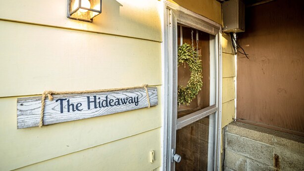 A wooden sign with "The Hideaway" written on it, attached to a building with a window and a door.