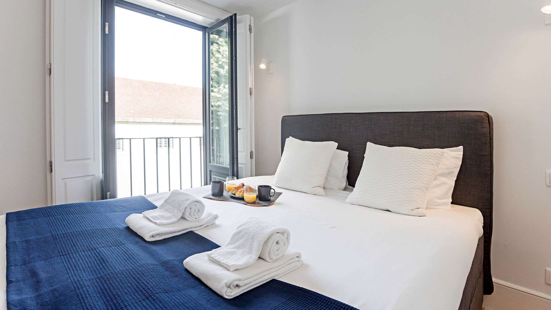 A hotel room with a bed, two pillows, a tray with breakfast items, and a window with a view of buildings.