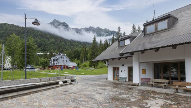 A building with a stone-tiled patio, a wooden bench, and a mountain backdrop. - Apartment in Braulins Near Tagliamento River (Chiusaforte)