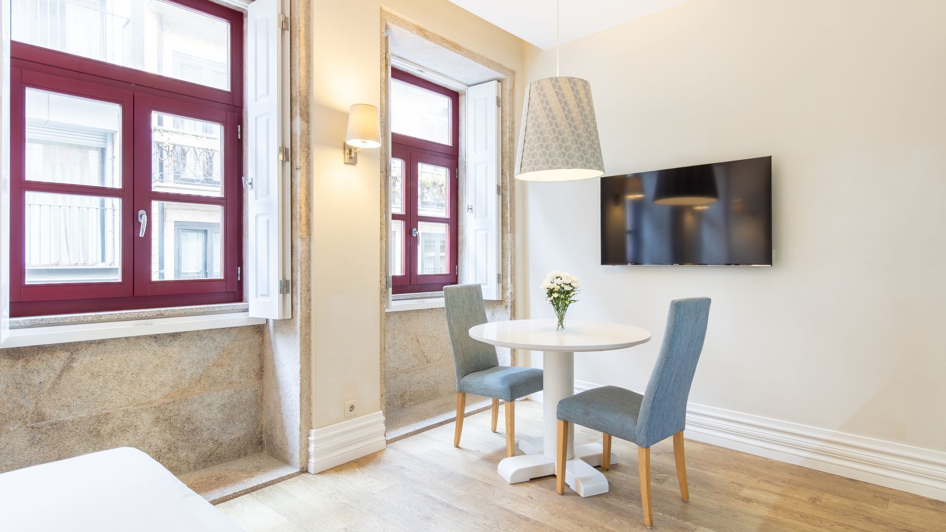 A room with a round table, two chairs, a TV mounted on the wall, and a door with red shutters.
