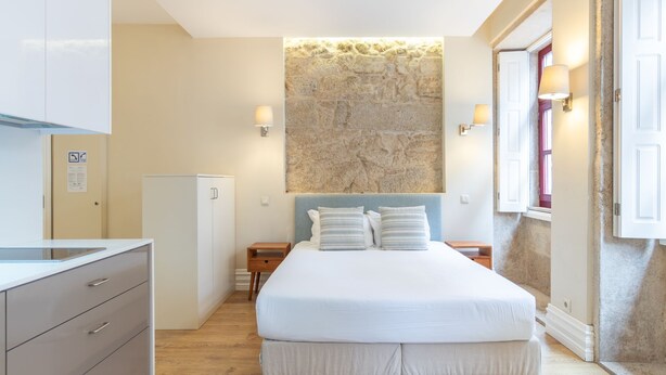 A bedroom with a bed, bedside tables, a wardrobe, a stone wall, and a window with a view. - Oporto Home Wine Cellars - Estúdio Standard - 1ºF (Vila Nova de Gaia)