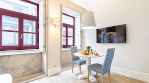 A dining area with a round table, two chairs, a TV mounted on the wall, and a window with red shutters.
