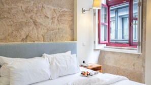 A bed with white linens, a stone wall, a window with red shutters, and a small wooden nightstand with a plate of berries. - Oporto Home Wine Cellars - Estúdio Standard - 1ºF (Vila Nova de Gaia)