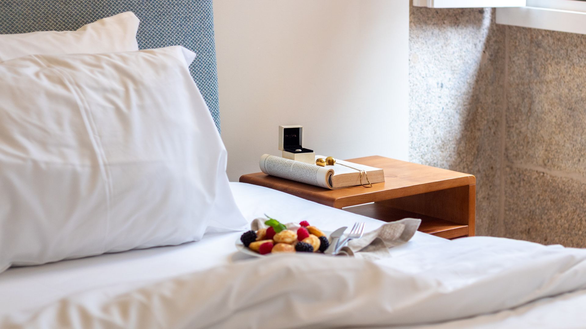 A neatly made bed with white linens, a small wooden bedside table with a book, a ring box, and a pair of earrings.