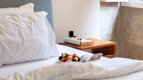 A neatly made bed with white linens, a small wooden bedside table with a book, a ring box, and a pair of earrings.
