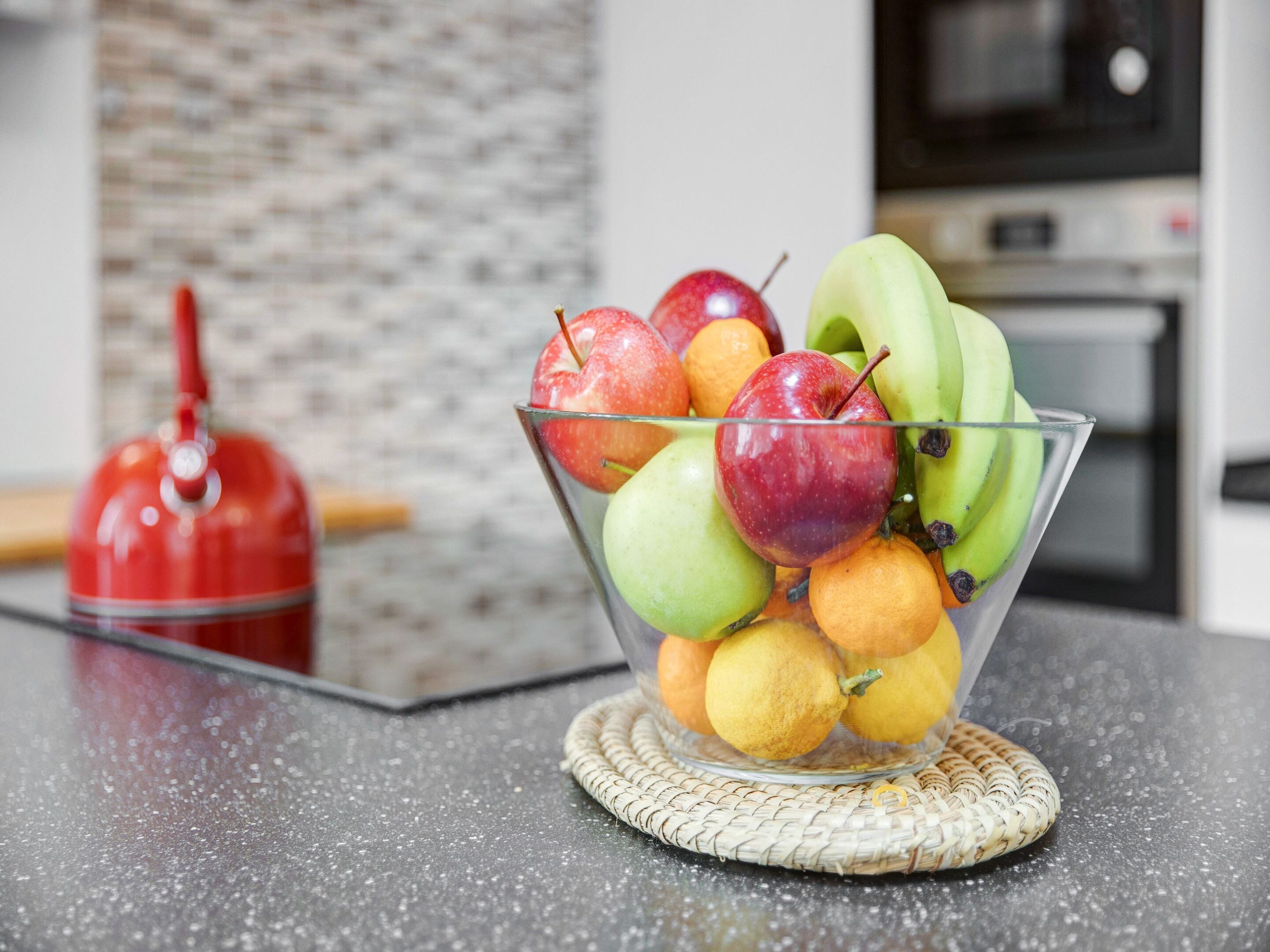 Uma tigela de frutas sobre uma bancada de cozinha, com uma chaleira vermelha ao fundo.