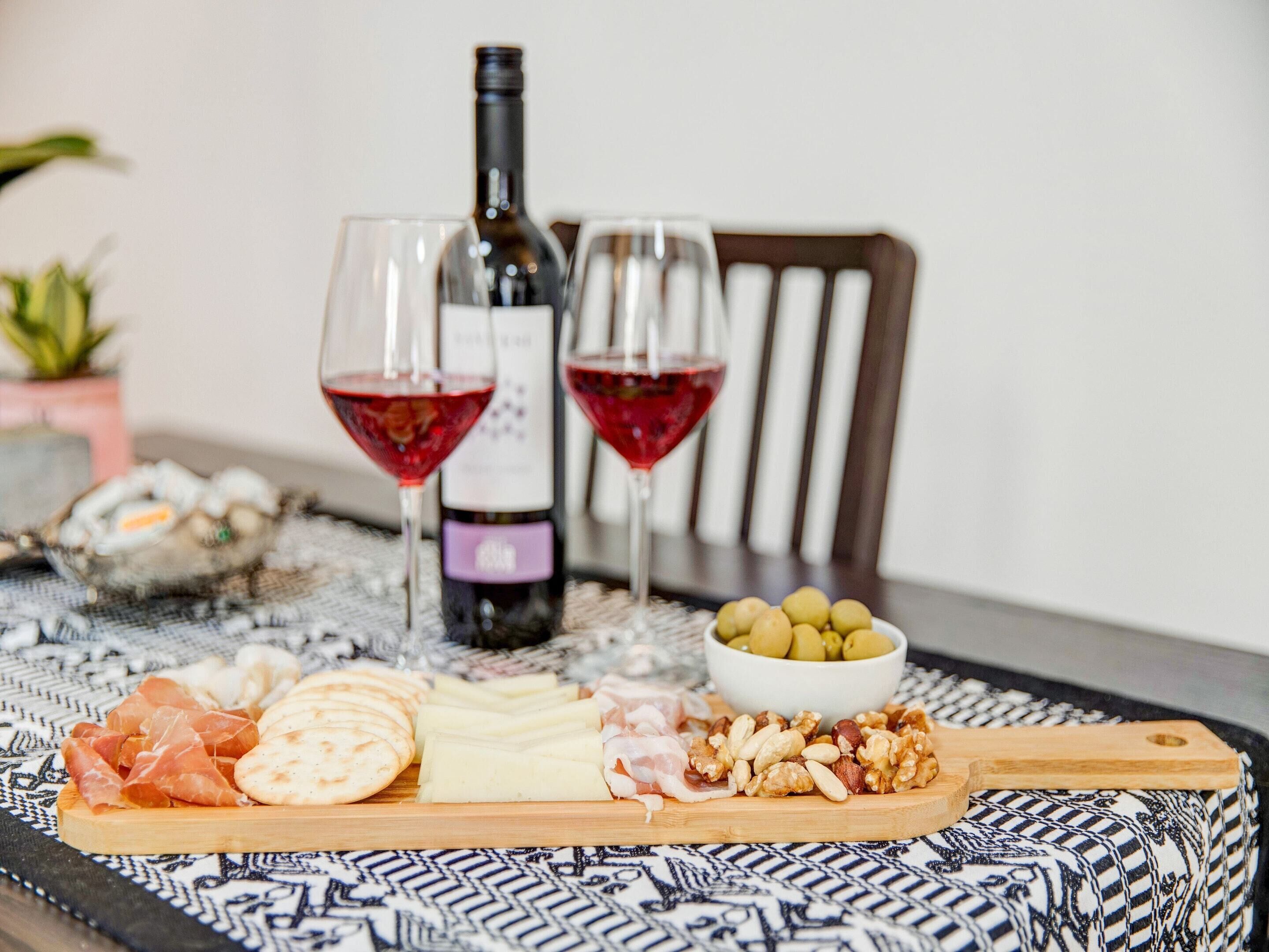 A table with a bottle of wine, two glasses, a bowl of olives, and a wooden board with various cheeses and meats.