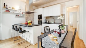 Una cocina moderna con armarios blancos, una isla central con taburetes de bar y una mesa de comedor para dos personas, con una bandeja de fruta y copas de vino.