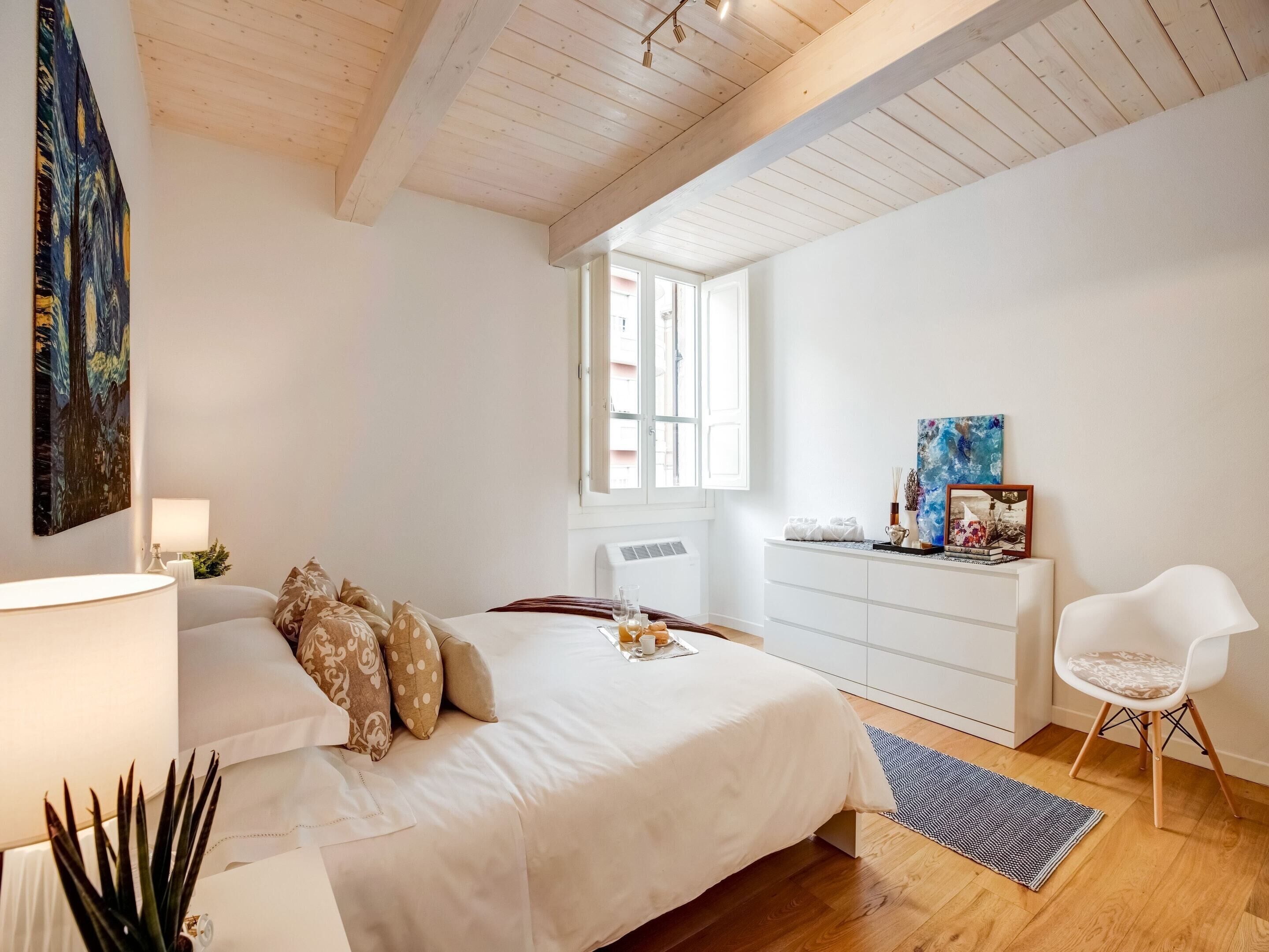 A bedroom with a bed, bedside table, lamp, dresser, chair, and artwork.