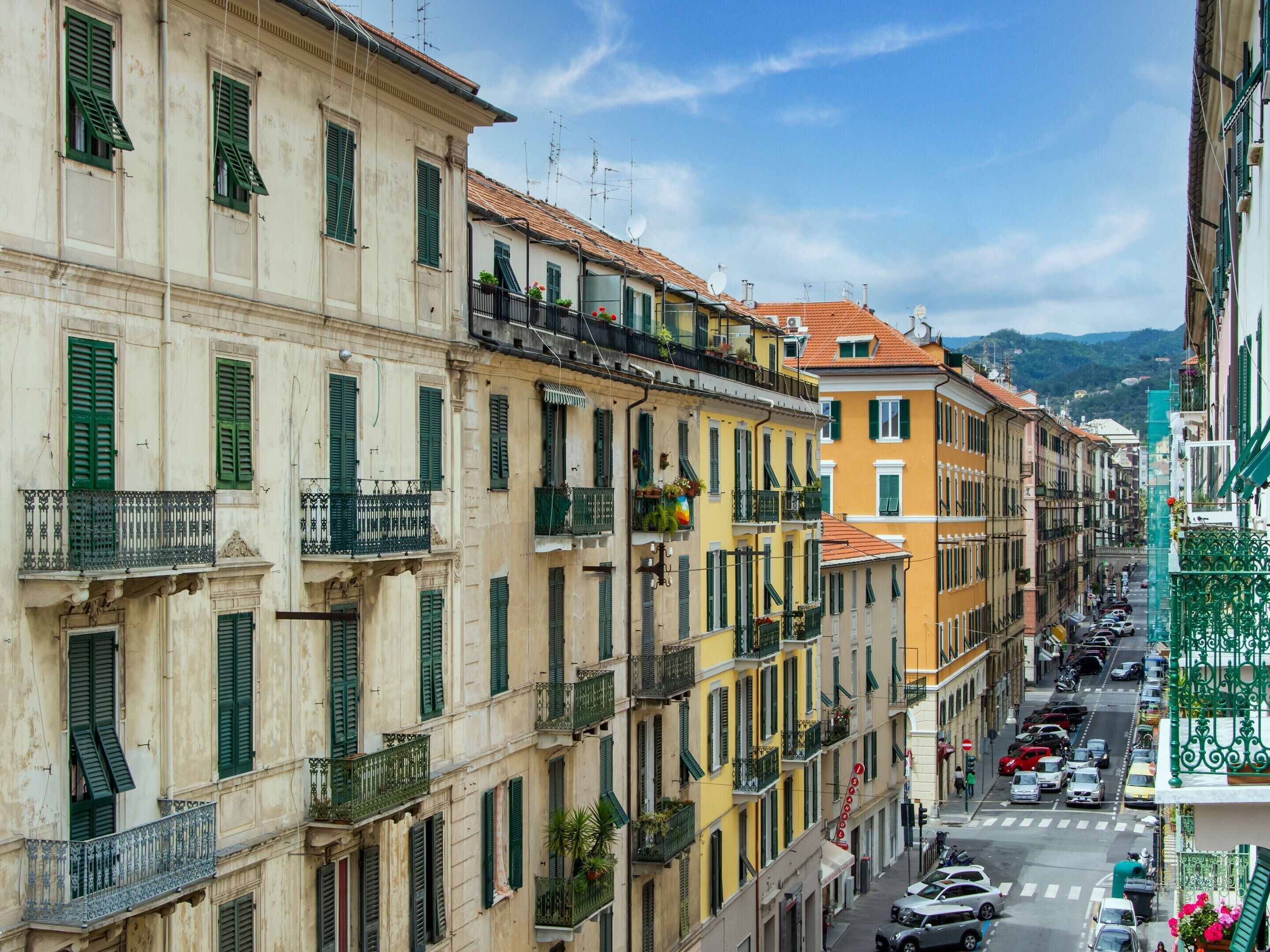 A row of multi-story buildings with balconies, a street with parked cars, and a clear sky.