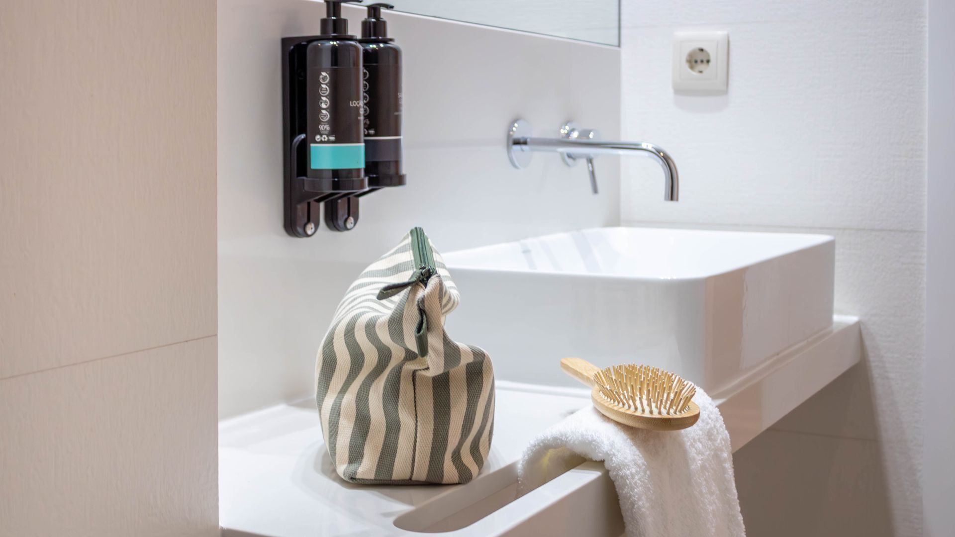 A bathroom with a white sink, a striped bag, a wooden brush, and two black bottles on the wall.