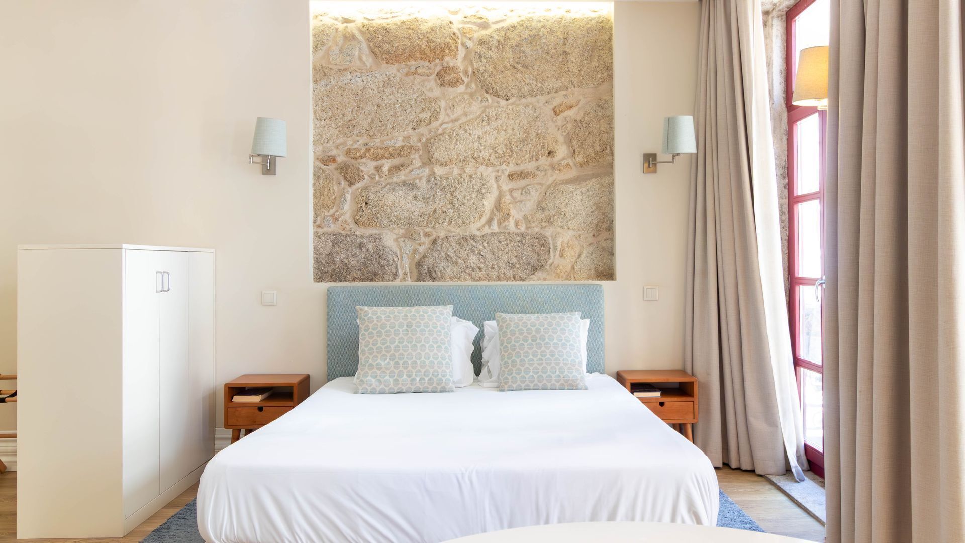 A bedroom with a stone wall, a bed with patterned pillows, bedside tables, and a wardrobe.