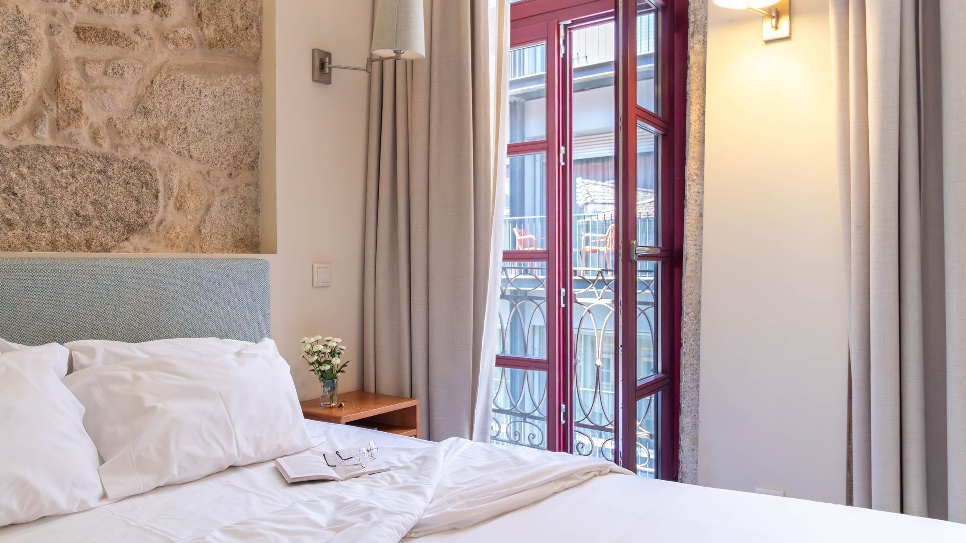 A hotel room with a stone wall, a bed with white linens, a nightstand with a vase of flowers, and a balcony with a red door.