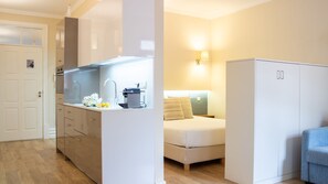 A compact hotel room with a kitchenette, a bed, a wardrobe, and a small seating area.