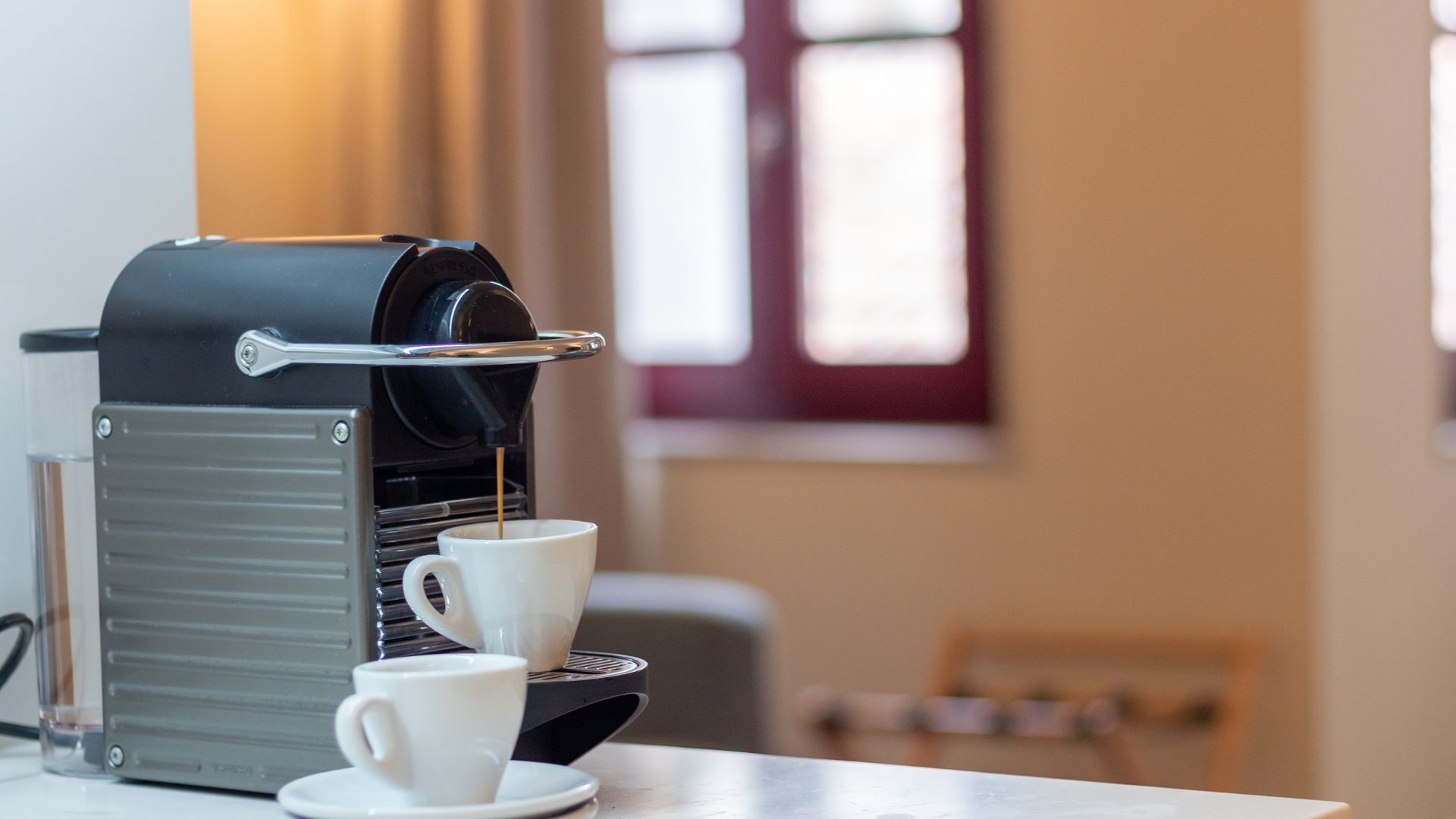 A coffee machine with two cups on a table, a window with blinds in the background.