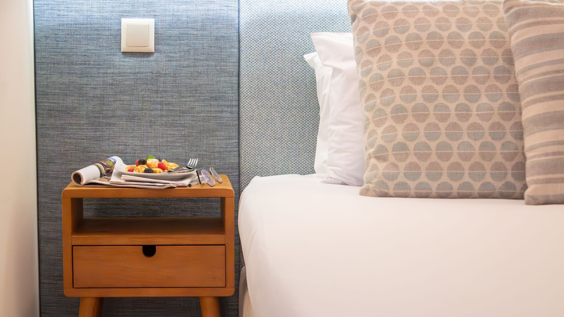 Een net opgemaakt bed met een gekleurd kussen, een houten nachtkastje met fruit en een krant, en een structuurmuur.