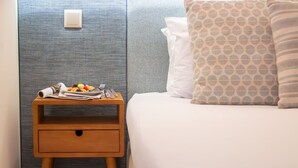 Un lit bien fait, agrémenté d’un coussin à motifs, une table de chevet en bois sur laquelle se trouvent une assiette de fruits et un journal, et un mur à la texture intéressante.