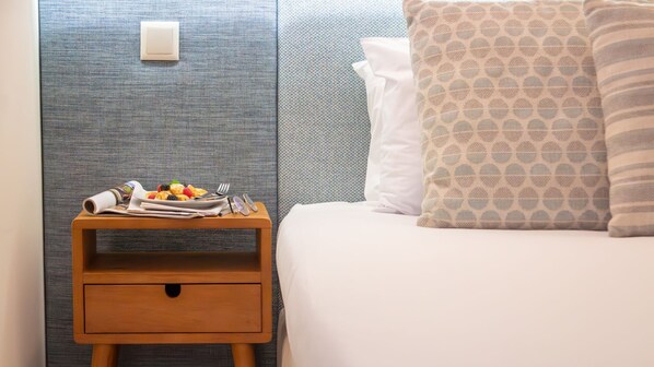A neatly made bed with a patterned pillow, a wooden nightstand with a plate of fruit and a newspaper, and a textured wall.