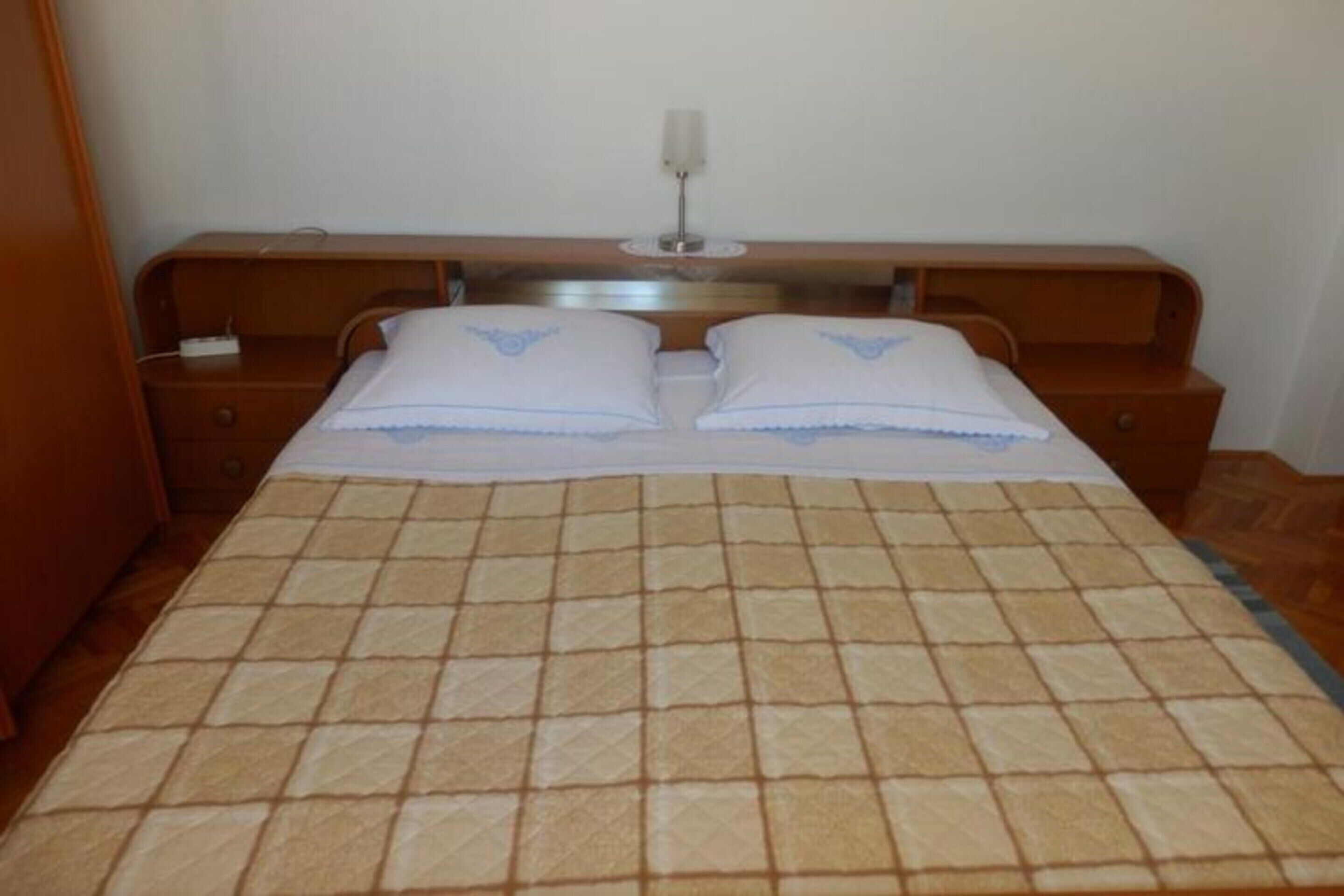 A double bed with a checkered bedspread, two pillows, a bedside table with a lamp, and a wooden nightstand.