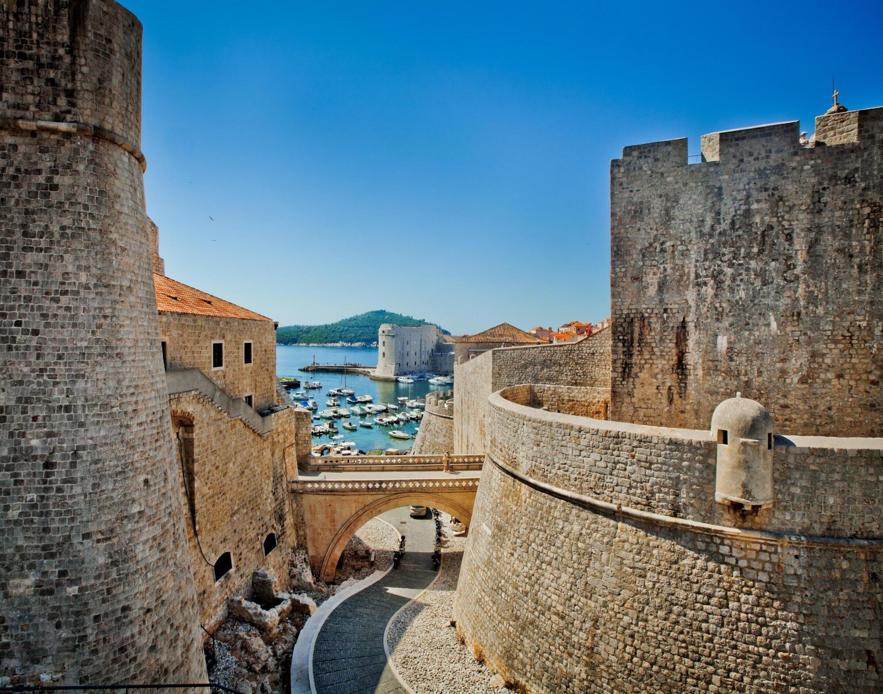 Una fortaleza de piedra histórica con un patio central, un puente y vistas a un puerto con barcos.