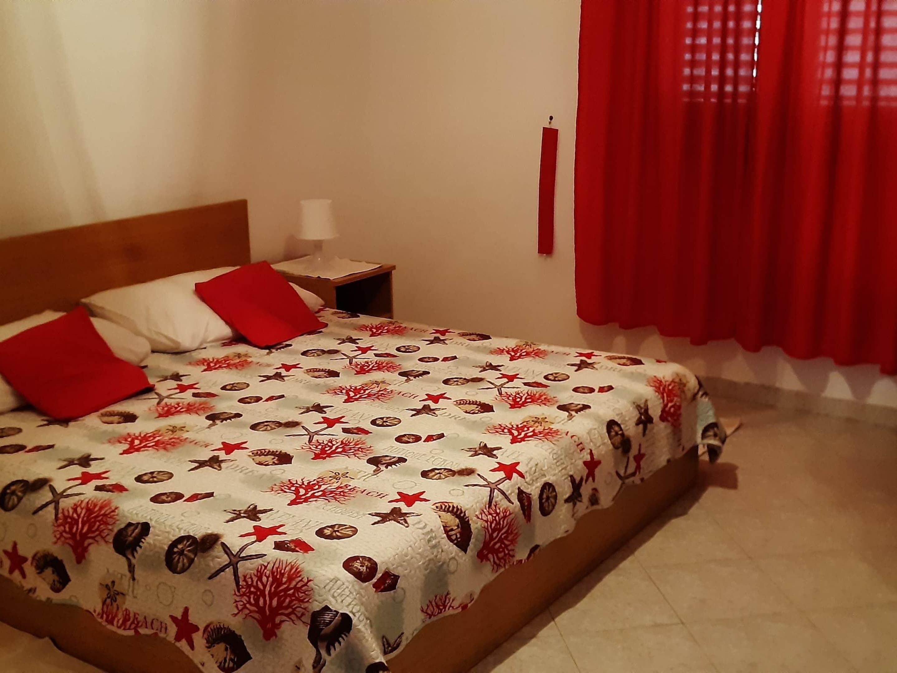 A neatly made bed with a patterned bedspread, a nightstand with a lamp, and red curtains.