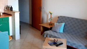 Una sala de estar con un sofá, una mesita con un control remoto y un libro, un florero con flores, y una zona de cocina con mostrador y armarios.