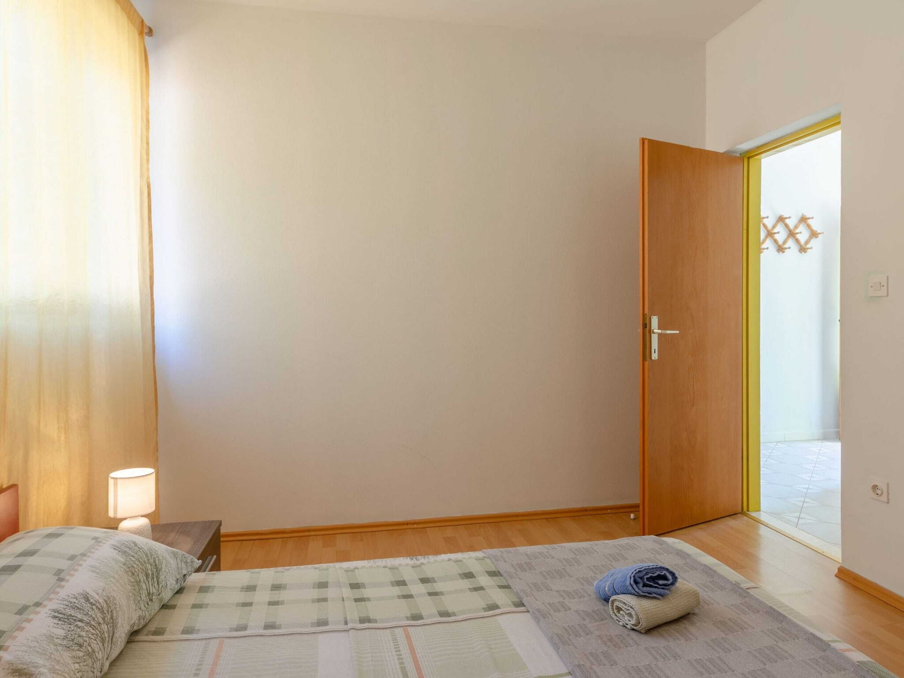 A bedroom with a bed, a bedside lamp, a wooden floor, a door with a yellow frame, and a view through the door.