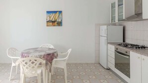Eine kompakte Küche mit Gasherd, Kühlschrank und weißen Schränken. Ein Esstisch mit Stühlen sowie ein Gemälde von Segelbooten an der Wand.