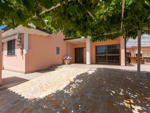 Ein Gebäude mit einer Steinterrasse, einem überdachten Eingangsbereich und einer Pergola mit Weinreben.