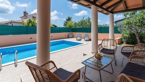 A covered outdoor pool area with lounge chairs and a glass table, surrounded by a green fence and trees. - Stylish Villa in Buje With Outdoor Kitchen (Buje)