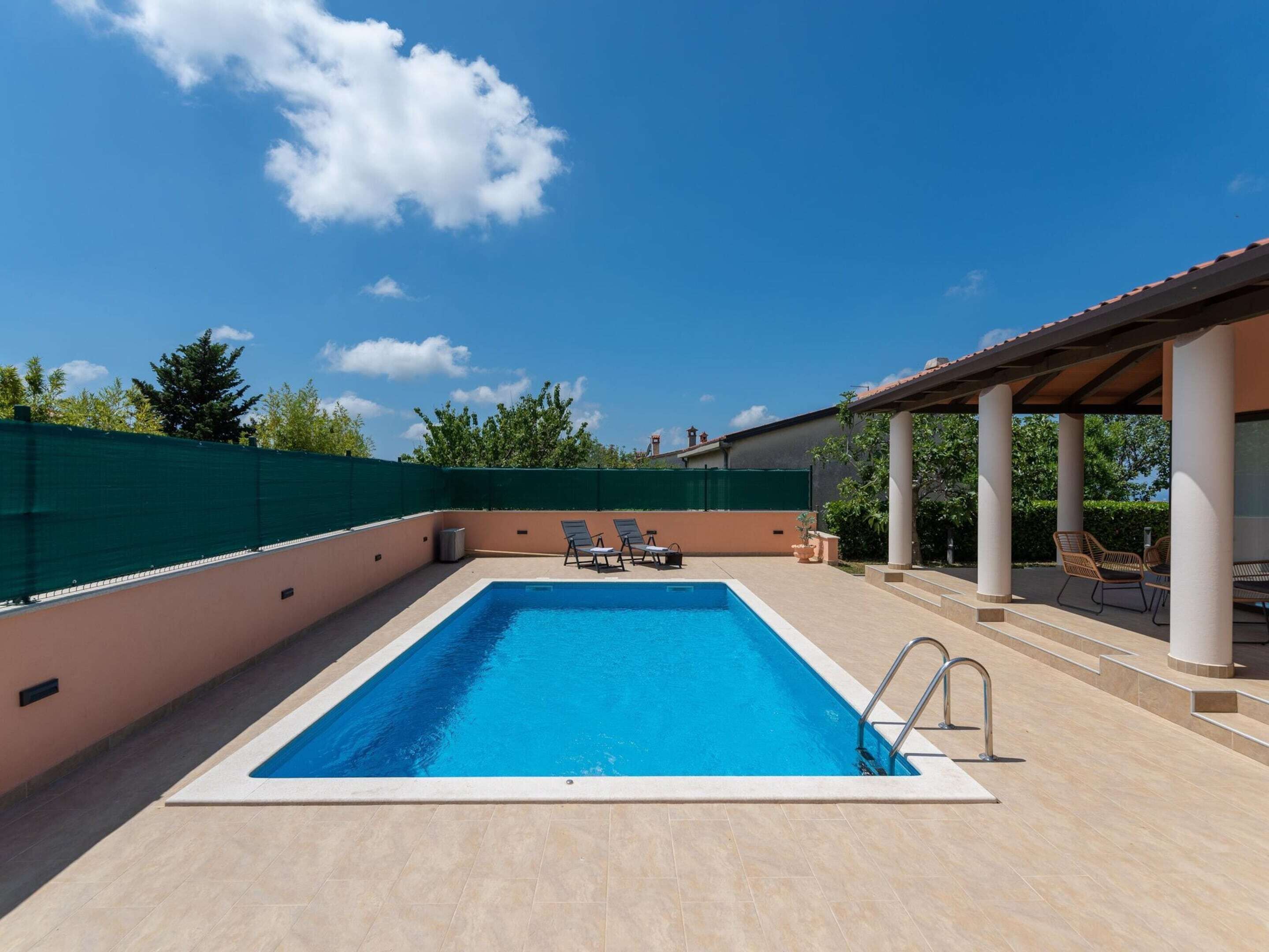 A rectangular swimming pool with a clear blue water, surrounded by beige tiles. There are two lounge chairs and a small table under a pergola.