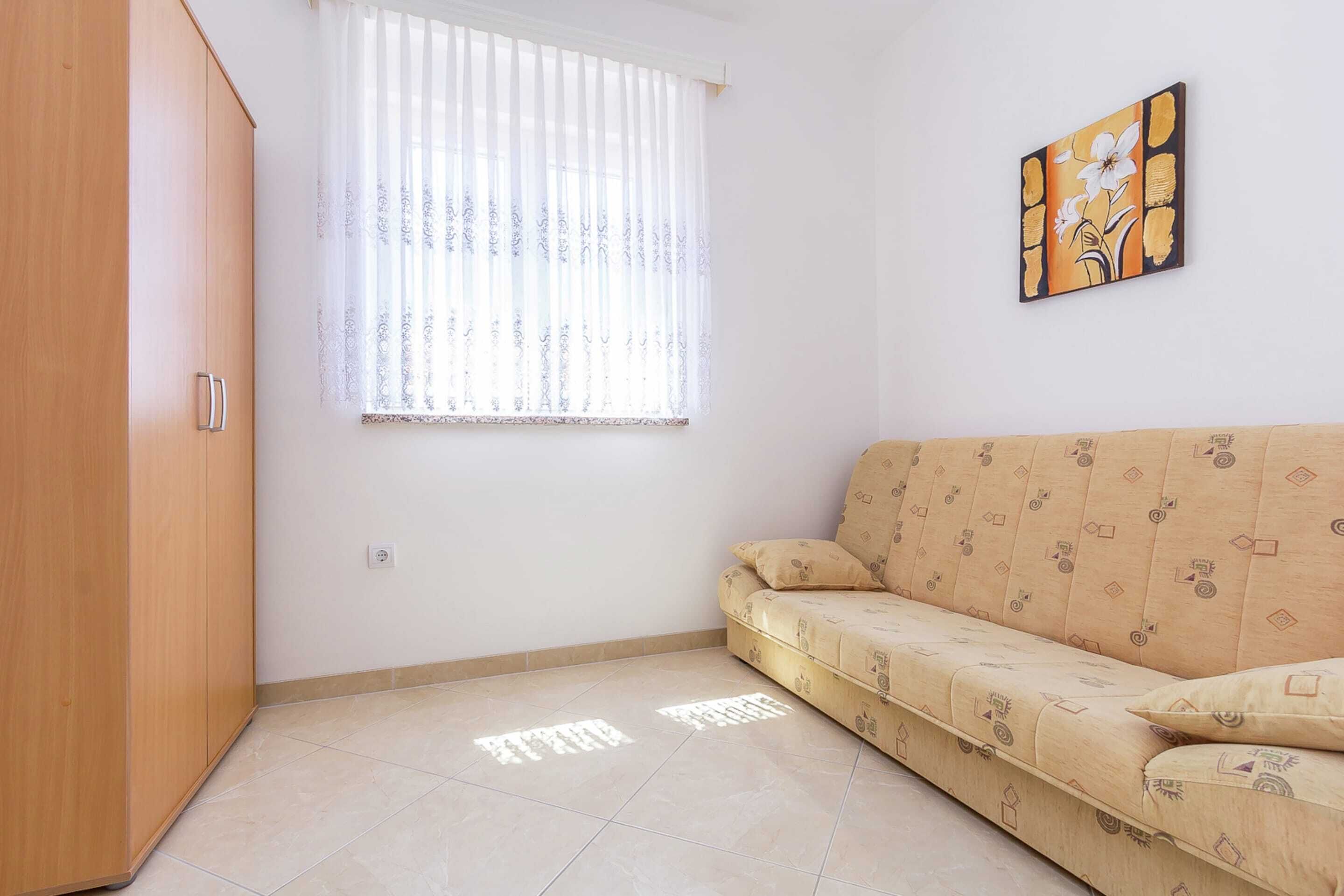 Una stanza con un divano color beige, un armadio in legno, una finestra con tende trasparenti e un quadro appeso al muro.