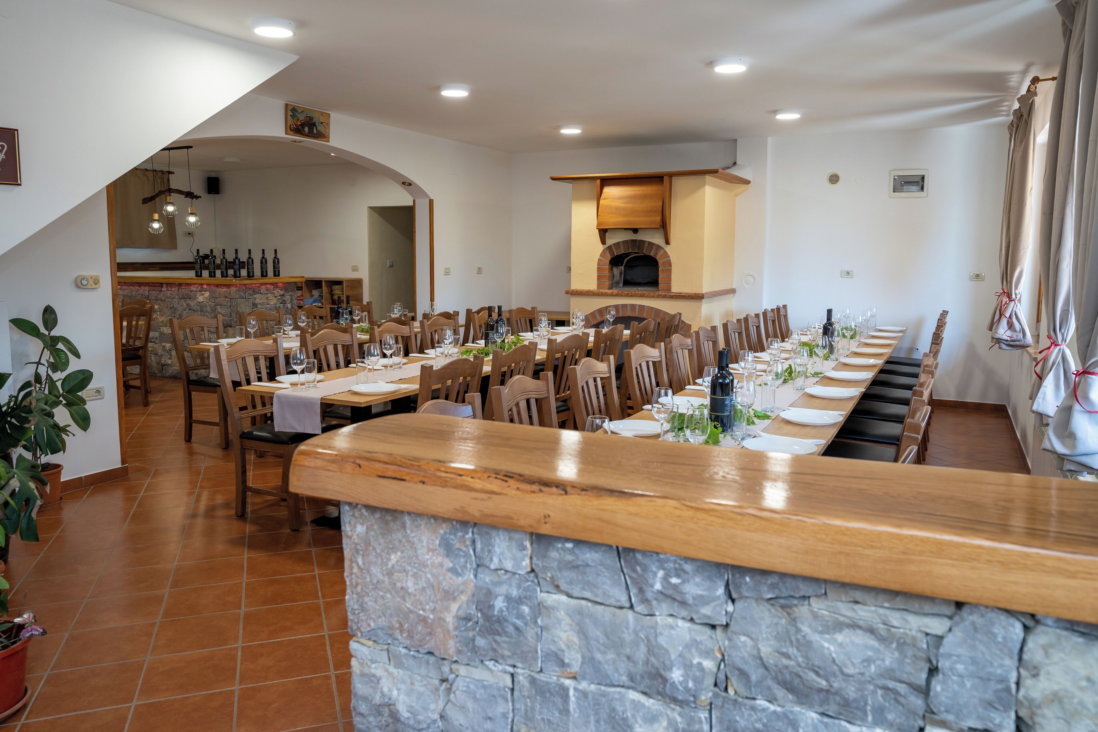 Un comedor amplio con una larga mesa de madera dispuesta para varios comensales, detalles de piedra y un horno de pizza empotrado.