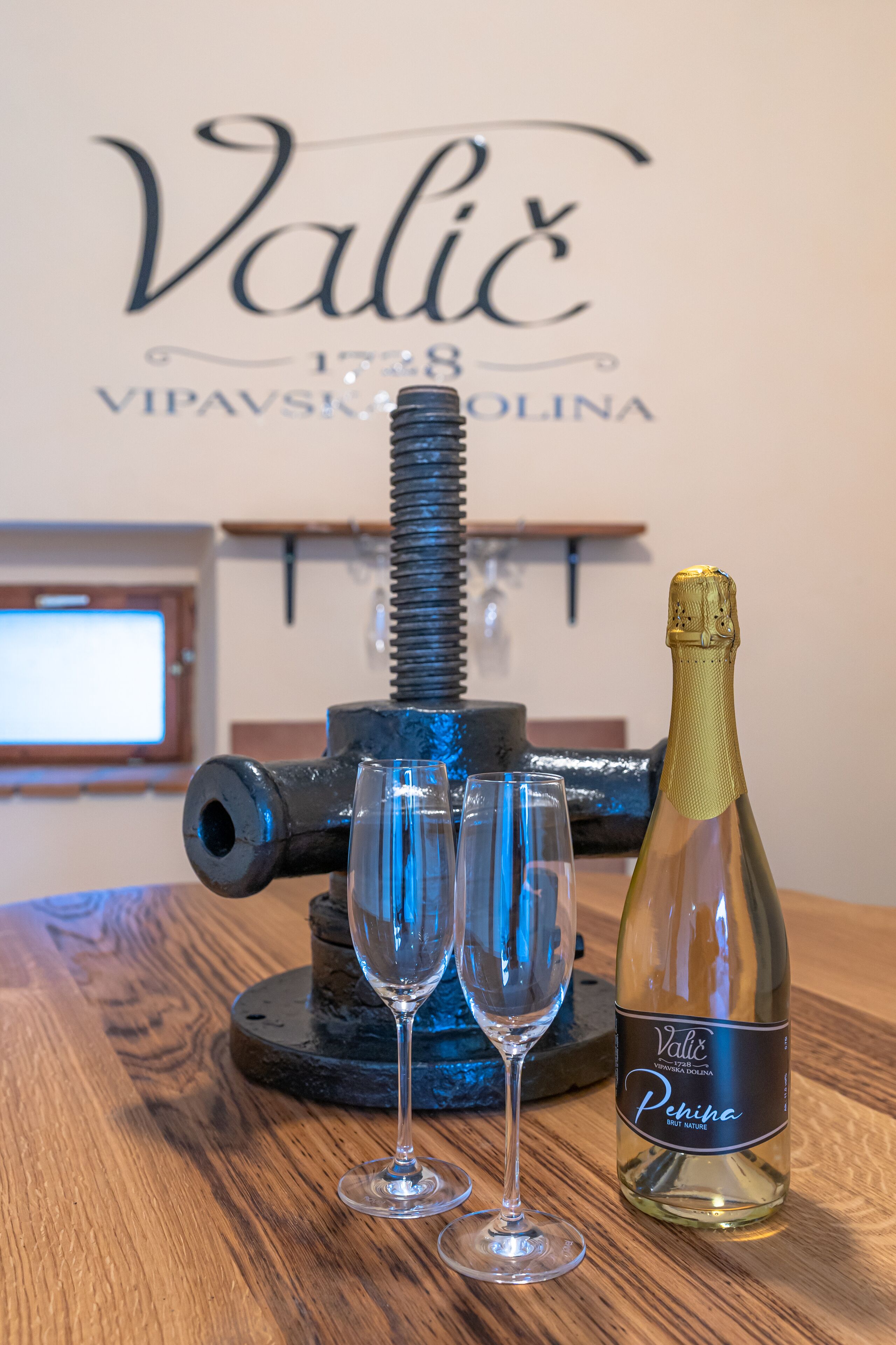A bottle of Valic Penina Brut Nature wine and two empty glasses on a wooden table, with a backdrop featuring the text "Valic 1828 VIPAVSKA OLINA".