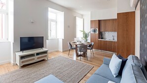Een moderne woonkamer met een flatscreen televisie, een grijze bank en een eethoek met een tafel en stoelen.
