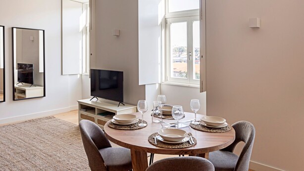 Een eetruimte met een ronde houten tafel gedekt voor vier personen, omringd door stoelen. Een televisie is aan de muur bevestigd, en er zijn twee ramen die voor daglicht zorgen.
