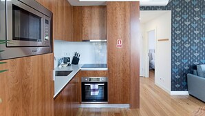 Een compacte keuken met een magnetron, broodrooster en oven, houten kastjes en behang met een patroon in de aangrenzende woonkamer.