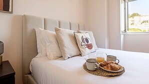 Een opgemaakt bed met kussens, een nachtkastje met lamp, en een ontbijt met croissants, koffie en sap.