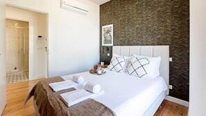 Une chambre d’hôtel avec un lit, une table de chevet avec une lampe, un tableau encadré au mur et une salle de bain visible à travers une porte ouverte.
