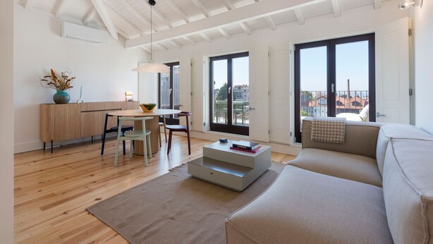 A modern living room with a sofa, a coffee table, a dining area with a table and chairs, and large windows offering a view of buildings outside.