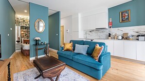 A modern living room with a teal sofa, wooden coffee table, and a kitchen area with white cabinets and a marble backsplash. - Turquoise Flat w/ AC & Balcony by LovelyStay (Porto)