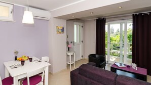 A modern living room with a white table, purple sofa, and a dining area with a white table and chairs. There is a large window with curtains, a door leading to a balcony, and a painting on the wall. - Apartments Gaura - Comfort One-bedroom Apartment With Terrace (Dubrovnik)
