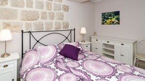 A bedroom with a stone wall, a bed with a patterned bedspread, a nightstand with a lamp, a dresser with drawers, and a chair. - Apartments Gaura - Comfort One-bedroom Apartment With Terrace (Dubrovnik)