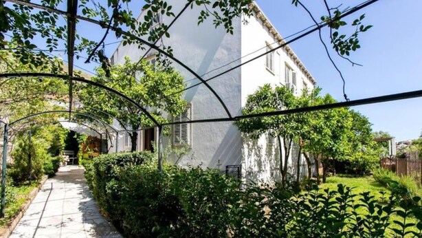 Un sendero conduce a un edificio blanco con contraventanas, rodeado de abundante vegetación y un cielo despejado.