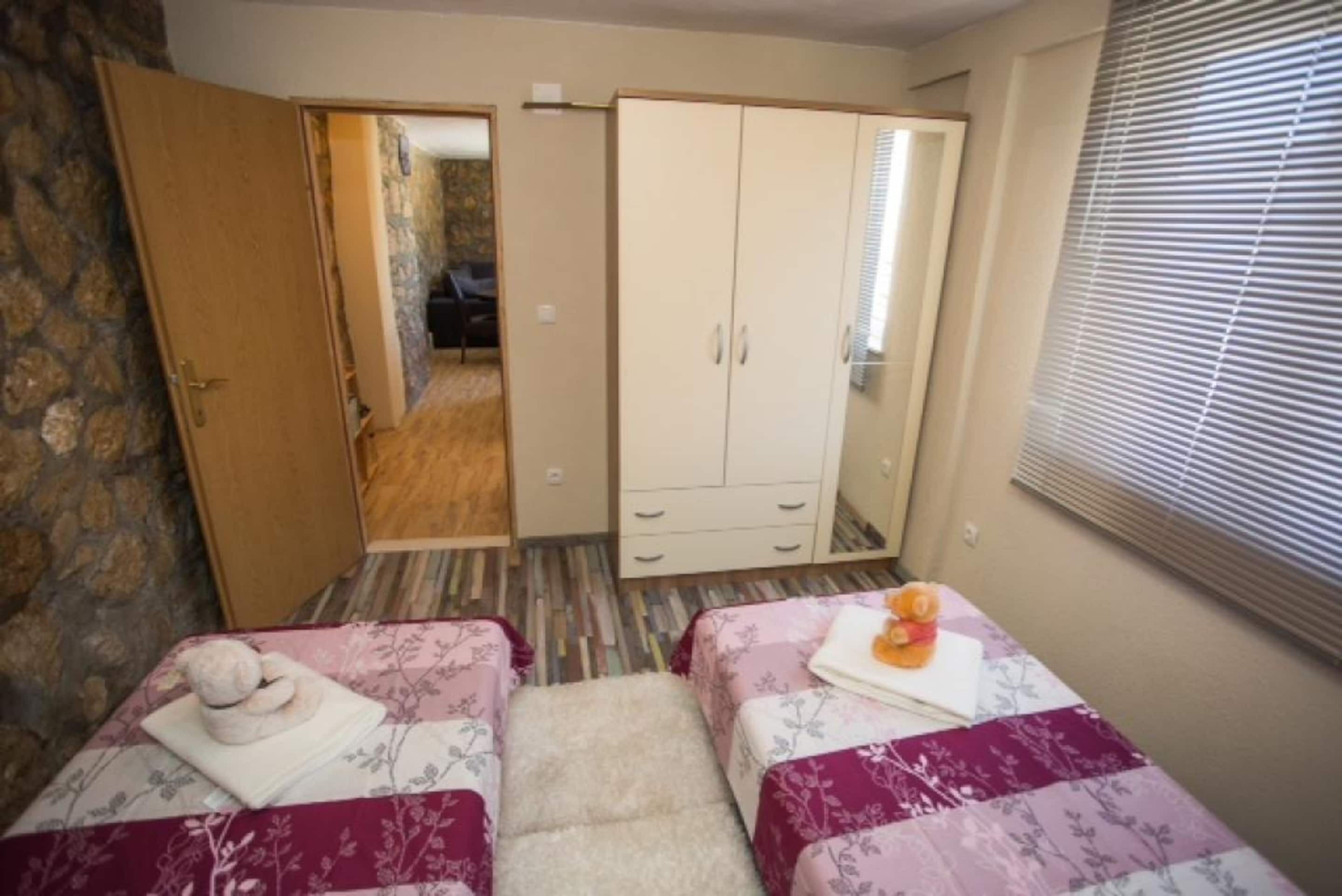 Une chambre avec deux lits, une armoire, un miroir et une fenêtre avec des stores.