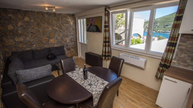 Ein gemütliches Zimmer mit Steinwand, einer Couch, einem Esstisch mit Stühlen, einer kleinen Küchenzeile und einem Fenster mit Meerblick.