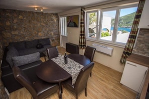 Una habitación acogedora con pared de piedra, un sofá, una mesa de comedor con sillas, una pequeña cocina y una ventana con vistas al mar.
