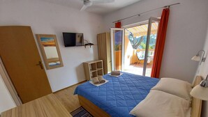 Ein Schlafzimmer mit einem Bett, einem an der Wand befestigten Fernseher, einem Holztisch, einer kleinen Kommode und einer Glastür, die zu einem Außenbereich führt.