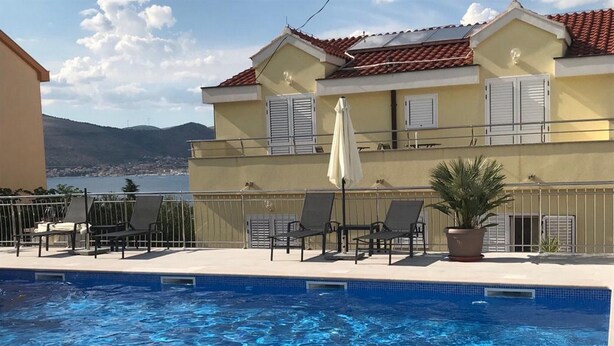 Une piscine avec des chaises longues et des parasols, un bâtiment avec un toit recouvert de tuiles rouges, et une vue sur la mer et les montagnes.
